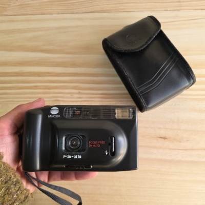 Câmara fotográfica Minolta FS-35 preta com flash e capa de proteção em pele preta sobre madeira clara.