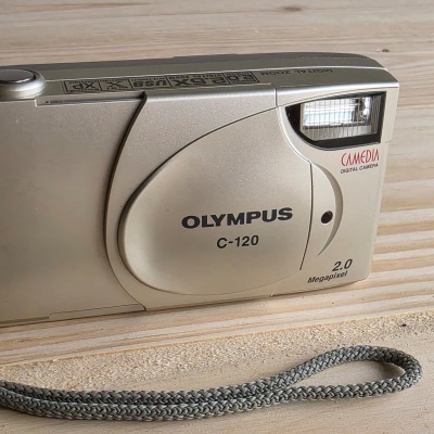 Câmara digital Olympus C-120 bege claro com flash e alça cinza sobre madeira clara