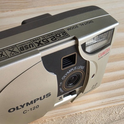 Câmara digital Olympus C-120 prata com lente e flash sobre mesa de madeira clara