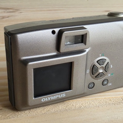 Câmara digital Olympus cinzenta com ecrã e botões num fundo de madeira.
