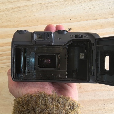 Câmara analógica preta com tampa aberta e mão segurando, sobre fundo de madeira clara