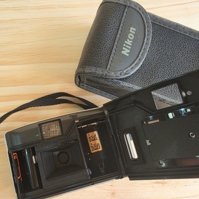 Máquina fotográfica analógica preta Nikon com capa preta aberta sobre superfície de madeira clara