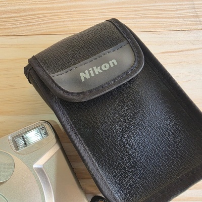 Capa preta com logo Nikon junto de parte de câmera prateada sobre madeira.