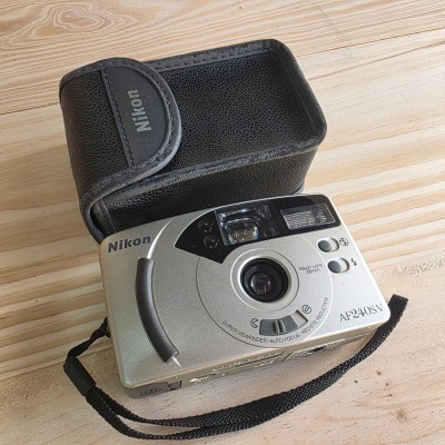 Câmara fotográfica compacta Nikon prata com bolsa preta sobre mesa de madeira.