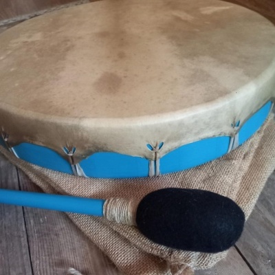 Tambor redondo com pele natural e batente azul sobre tecido rústico e chão de madeira