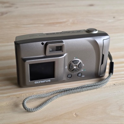 Máquina fotográfica digital Olympus cinza com ecrã LCD e pulseira cinzenta