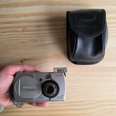 Câmara digital Olympus prateada com flash aberto e bolsa preta sobre madeira clara