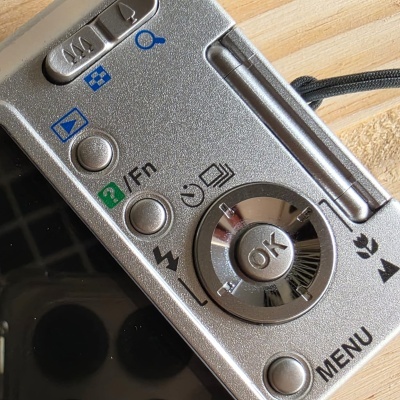 Painel de controlo prateado de câmara digital com botões metálicos e texto ?/Fn e MENU.