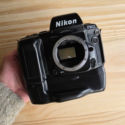 Máquina fotográfica Nikon F90X preta sem lente, segurada à mão sobre superfície de madeira.