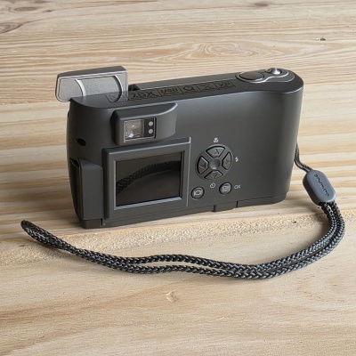 Câmara fotográfica compacta cinzenta com ecrã e flash aberta, sobre mesa de madeira.