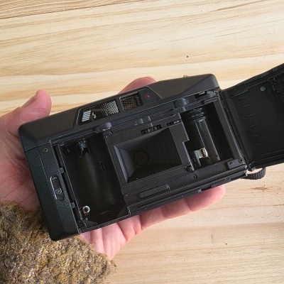 Câmara fotográfica analógica preta aberta sobre uma mão e fundo de madeira clara