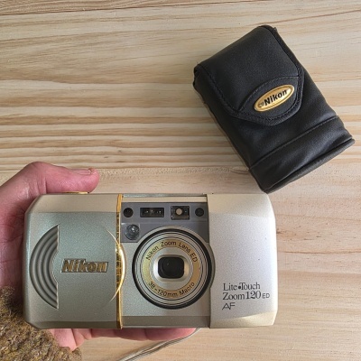 Câmara fotográfica compacta Nikon prateada com detalhes dourados e estojo preto.