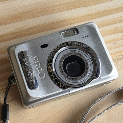 Câmara digital prateada PENTAX Optio com lente e flash, cordão e superfície de madeira clara