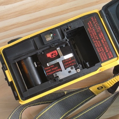 Máquina fotográfica analógica amarela com interior preto aberto e alça de tecido preta com detalhe amarelo
