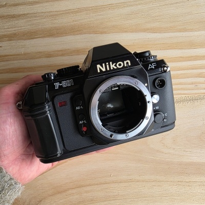 Máquina fotográfica analógica Nikon F-501 preta sem lente numa mão sobre superfície de madeira