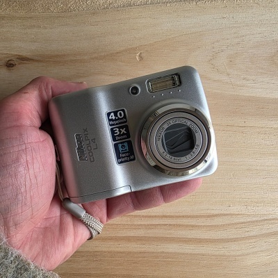 Câmara digital compacta prateada Nikon COOLPIX L4 vista de frente numa mão com fundo de madeira.