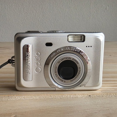 Câmara fotográfica digital prateada Pentax Optio com lente circular e flash embutido sobre superfície de madeira clara