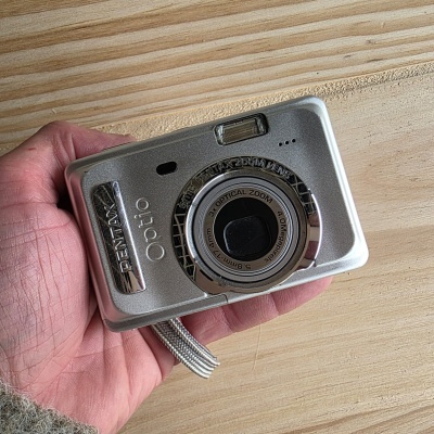 Câmara digital Pentax Optio prata com lente e flash, mão segurando, fundo de madeira