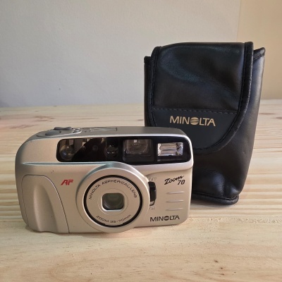Câmara Minolta Zoom 70 prateada com capa preta sobre mesa de madeira.
