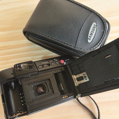 Câmara fotográfica analógica preta aberta com estojo preto Samsung sobre mesa de madeira clara