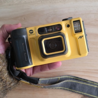 Máquina fotográfica analógica amarela e preta MINOLTA com flash integrado e alça cinza com detalhes amarelos.