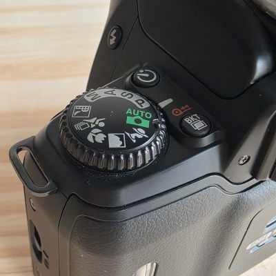 Câmara fotográfica preta com roda de modos e botões visíveis