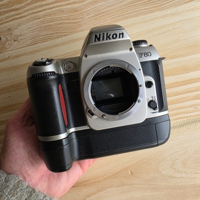 Câmara fotográfica analógica Nikon F80 preta e cinza numa mão sobre madeira clara
