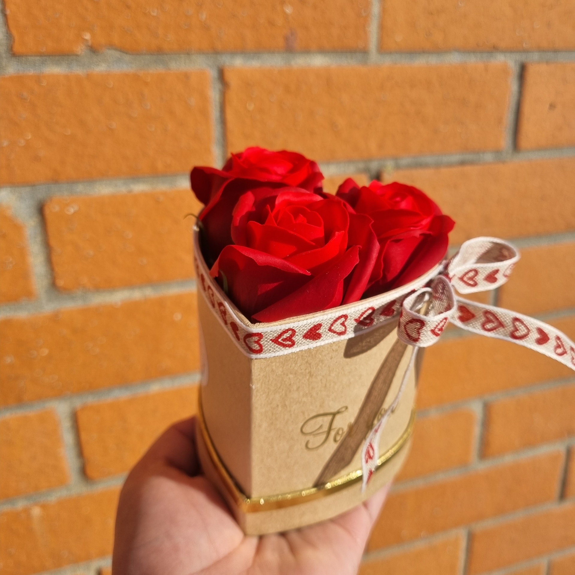 Caixa pequena com três rosas vermelhas e fita com corações vermelhos contra parede de tijolos.