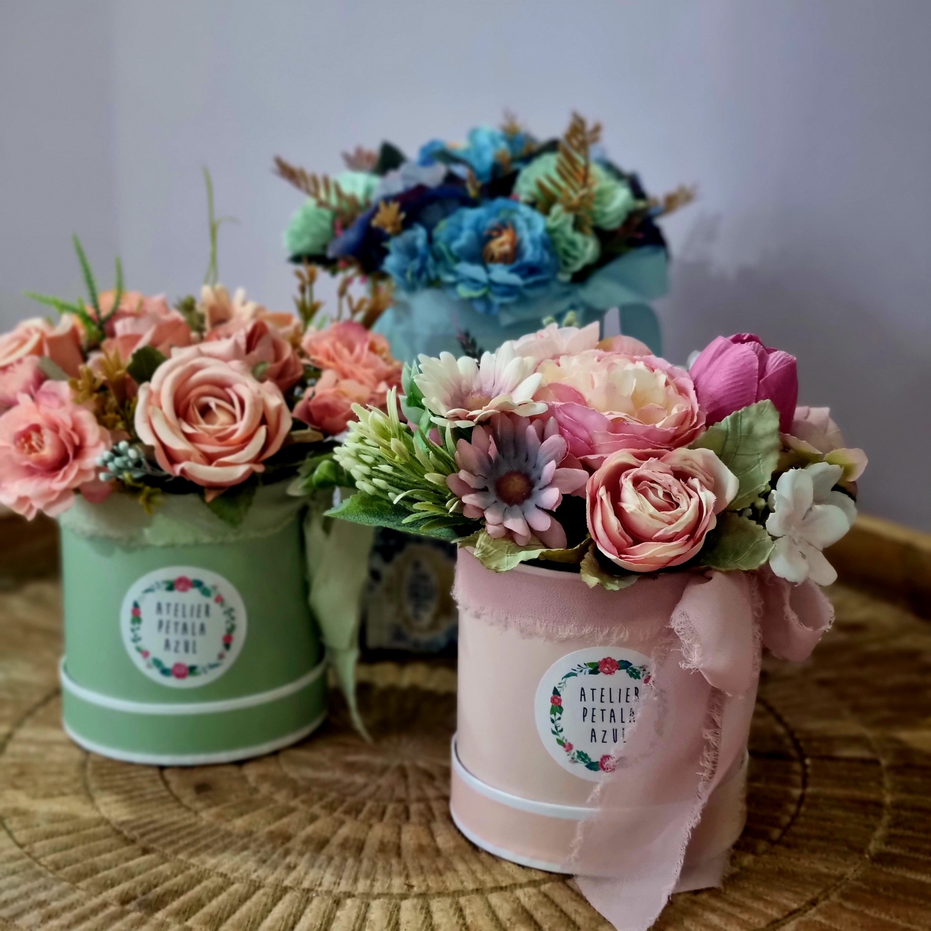 Três arranjos florais coloridos em vasos redondos decorativos com etiqueta ATELIER PETALAS AZUL sobre mesa de madeira