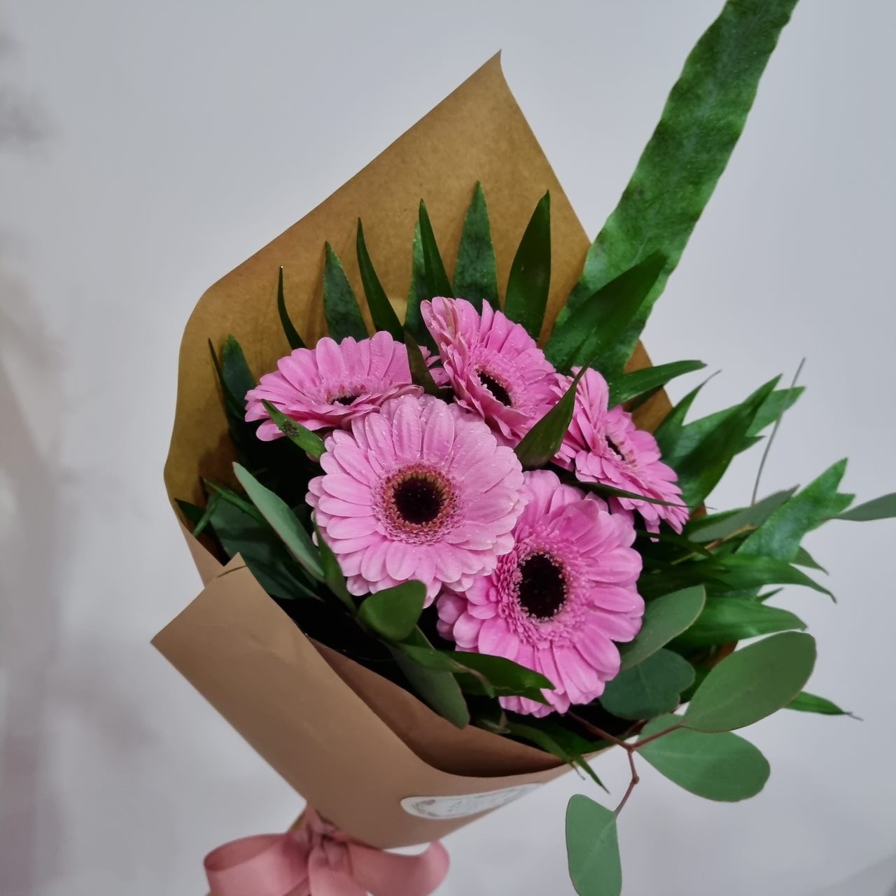 Ramo de gerberas cor-de-rosa com folhas verdes, embrulhado em papel kraft e fita rosa