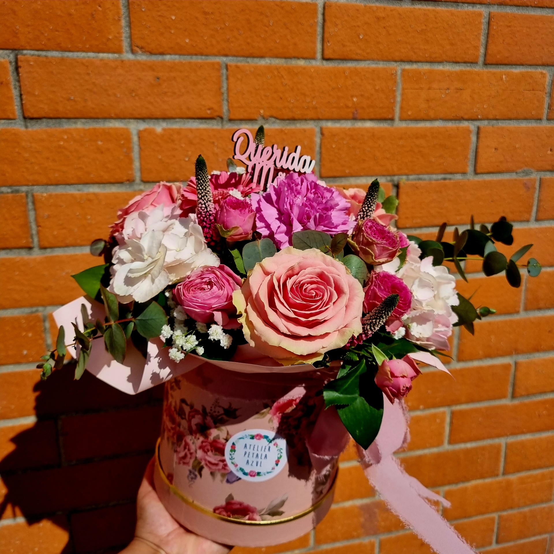 Arranjo floral em caixa redonda decorada com padrão de rosas, com flores rosa, cravos, folhagem verde e adorno de madeira 