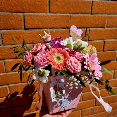 Arranjo de flores coloridas num vaso rosa com placa de coração e cartão Dia da Mãe segurado à frente de parede de tijolos.