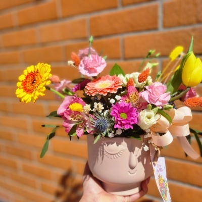Vaso de cerâmica rosa com rosto feminino e flores coloridas, etiqueta 'Dia da Mãe', parede de tijolos ao fundo