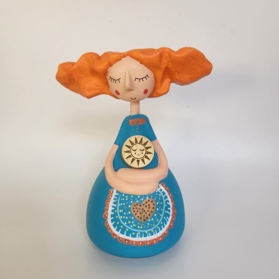 Figura artesanal de mulher com cabelo laranja ondulado, vestido azul e disco dourado com sol sorridente