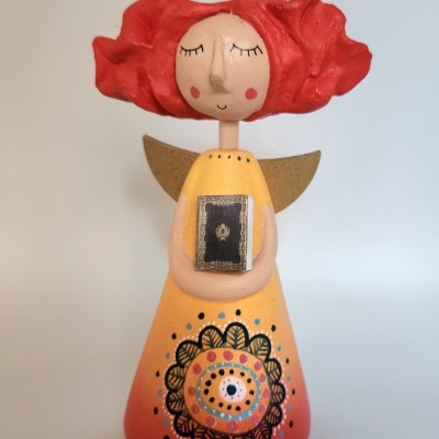 Escultura de anjo com vestido amarelo e cabelo vermelho segurando livro