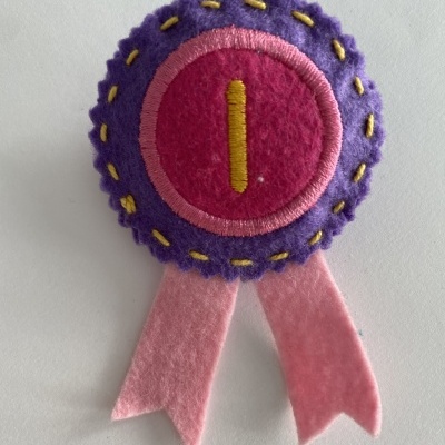 Medalha de feltro roxa e rosa com detalhe amarelo e duas fitas rosa