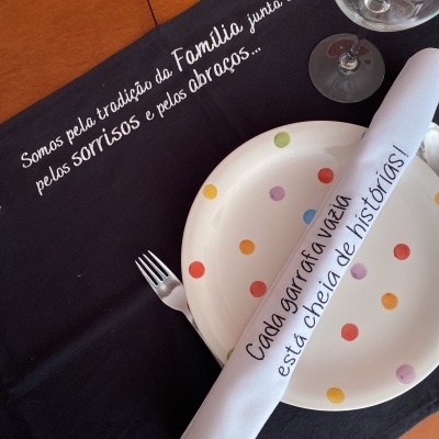Mesa posta com prato branco com bolinhas coloridas, guardanapo branco com texto em português, garfo, copo e toalha preta com texto em branco
