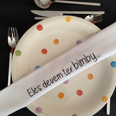 Mesa posta com prato branco com bolinhas coloridas e talheres em pano preto com texto bordado