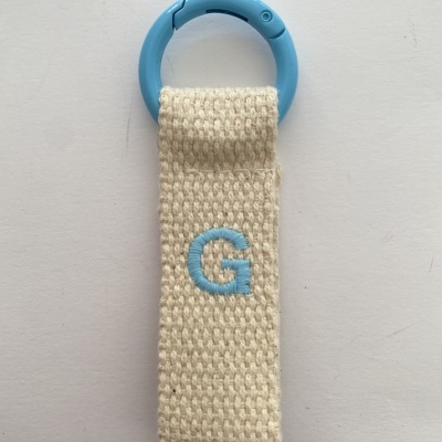 Porta-chaves com argola azul e fita de tecido bege com letra G bordada.