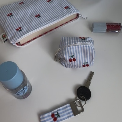 Bolsa, porta-moedas e porta-chaves de tecido azul listrado com cerejas bordadas, chave, batom Wet Lip Oil, frasco azul e objeto decorativo sorridente sobre mesa branca.