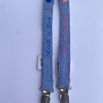 Clips para chupeta em tecido azul claro com fecho metálico e textos 'I'm a boy' e 'I'm a girl'.