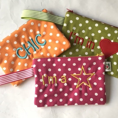 Três bolsas pequenas com padrão de bolinhas e frases bordadas, em tecido colorido.