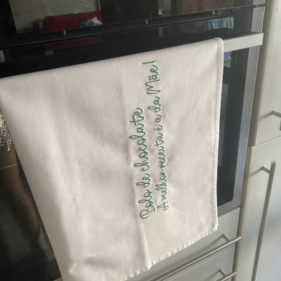 Toalha de cozinha branca bordada em verde pendurada na maçaneta de um forno inox.