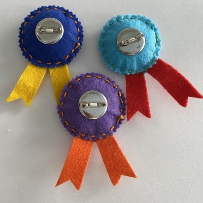 Três broches circulares de feltro colorido com fitas e bordados, vistos pelo verso com fechos metálicos.