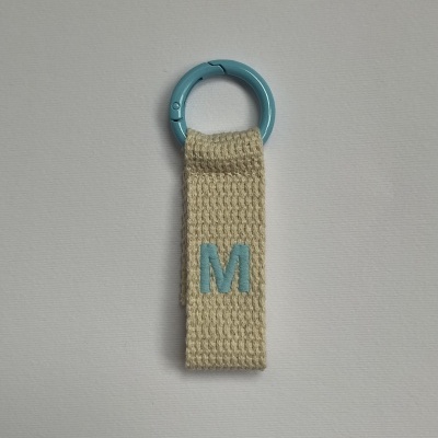 Chaveiro com argola azul e fita de tecido bege com letra M.