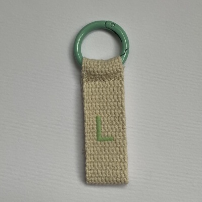 Porta-chaves com argola verde pastel e tira de tecido bege com letra L bordada