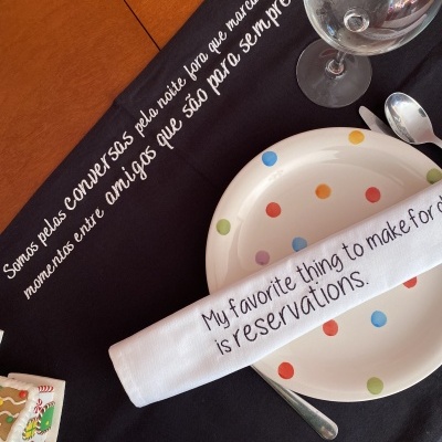 Mesa posta com toalha preta com texto em português, prato com bolinhas coloridas, guardanapo branco com texto em inglês e copo de vidro