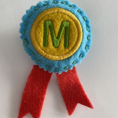 Emblema de feltro azul, amarelo e vermelho com letra M verde