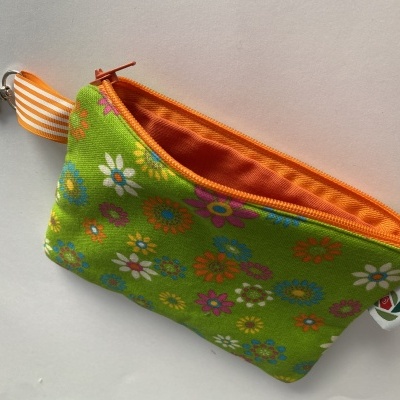 Carteira pequena verde com padrão de flores coloridas, interior laranja, com laço laranja às riscas e mosquetão metálico