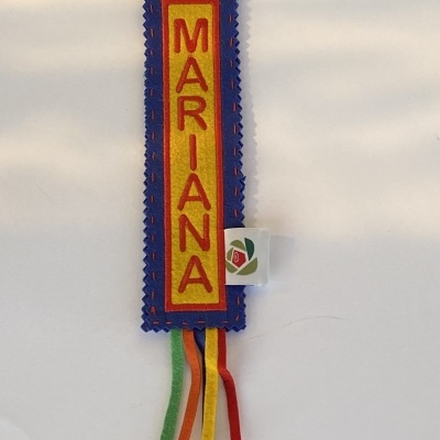 Marcador de livro tecido com nome MARIANA em letras vermelhas sobre fundo amarelo com bordas azuis e fios coloridos pendurados.